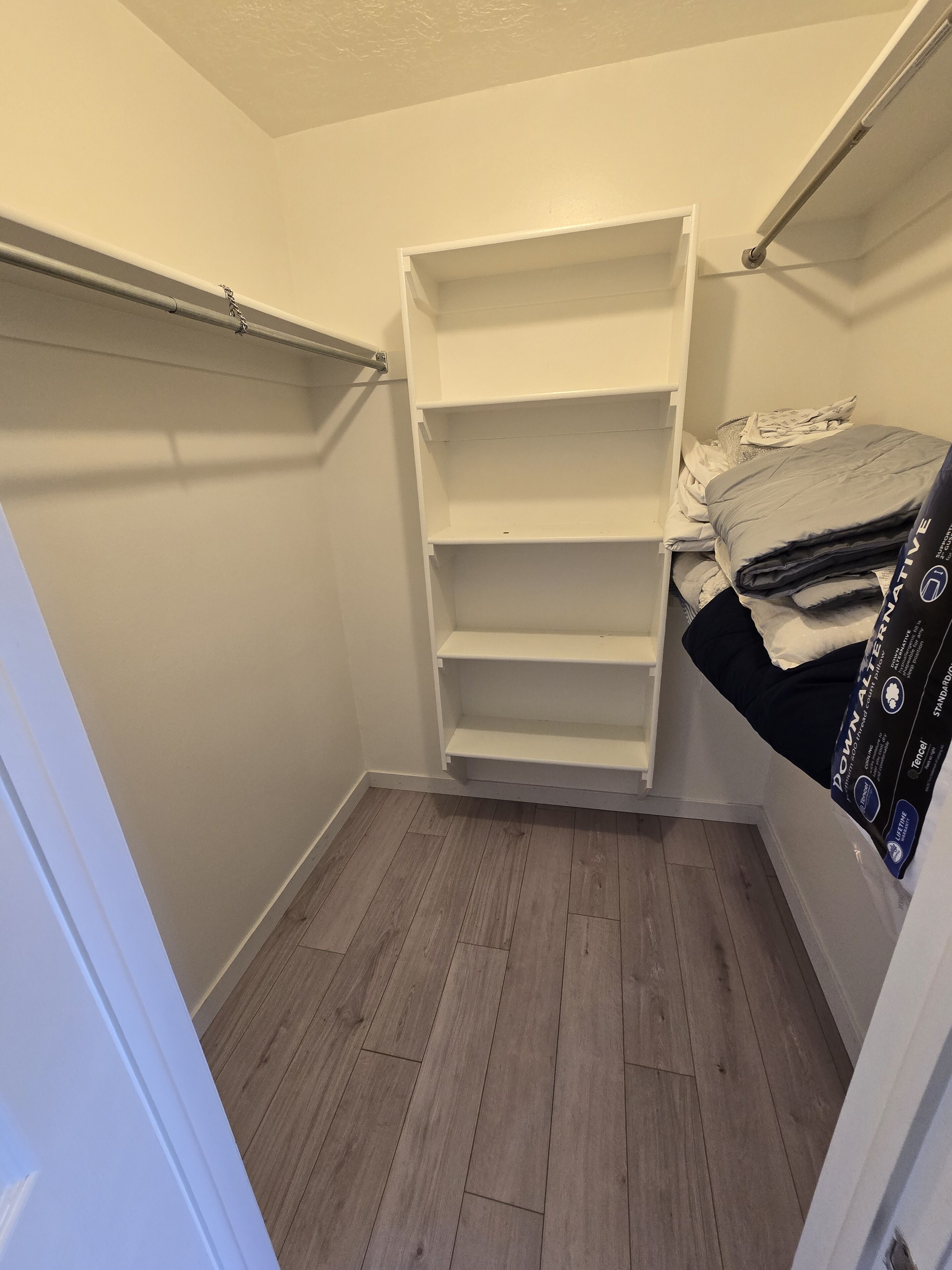 Een kleine, smalle kamer met een houten vloer en witte wanden. Er is een ingebouwde boekenkast met meerdere planken en een bed met een zwarte deken en een kussen.