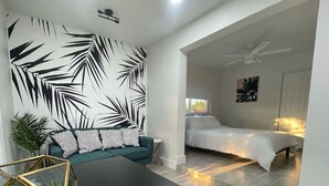 A modern living room with a black and white leaf-patterned wall, a teal sofa, a glass coffee table, and a bed with a white duvet in the adjacent room. - The Vice City Suite•Private Stay+Free Parking in the Heart of Miami (Miami)
