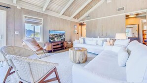 A spacious living room with a high ceiling, a large flat-screen TV, a white sofa, and a wicker chair.