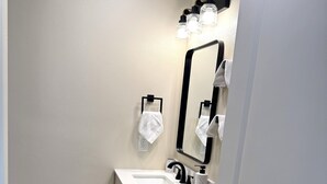 A bathroom with a white sink, black faucet, and a mirror with lights above it. There are two towels hanging on hooks and a soap dispenser on the sink.