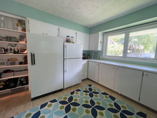 A kitchen with white cabinets, a refrigerator, a window, and a patterned rug. - Stunning 4-Bed, 3-Bath with 100 Ft of Private Beach – Your Dream Retreat Awaits (Traverse City)