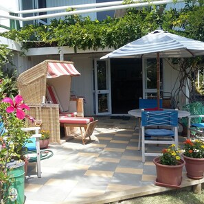 Een terras buiten met een tafel, stoelen, een rieten stoel met een gestreepte parasol en potplanten.