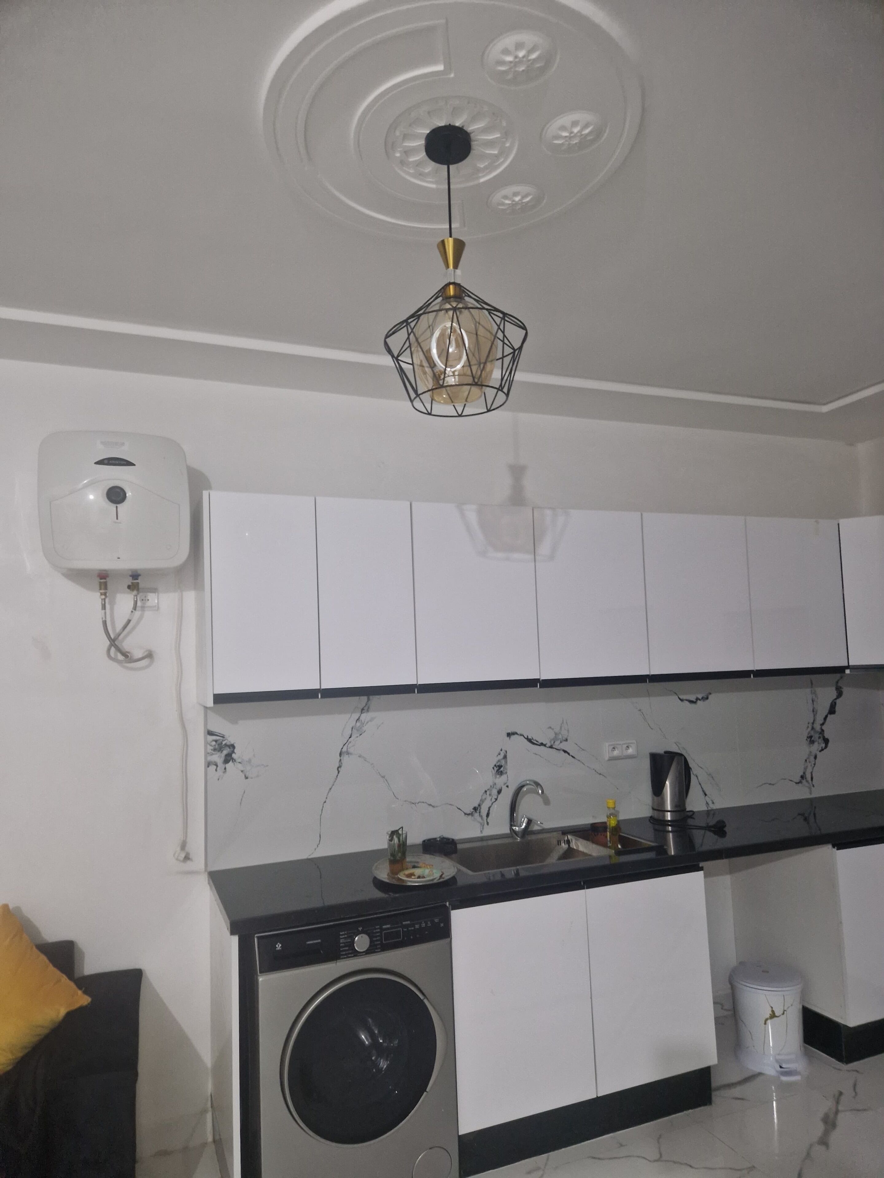 Une cuisine compacte avec des meubles blancs, un pare-feu en marbre, un lave-linge et un luminaire au plafond.