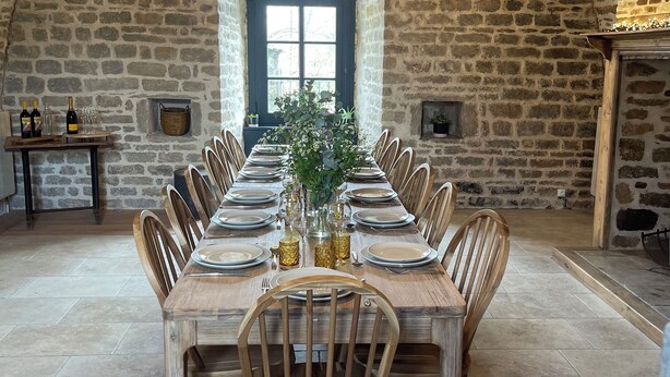 Una larga mesa de comedor de madera dispuesta para una comida en una habitación de paredes de piedra con techo alto y un gran ventanal.