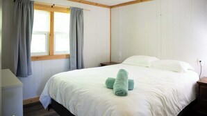 A neatly made bed with white linens and two rolled towels, a wooden nightstand, a window with curtains, and a white wall with wooden trim.