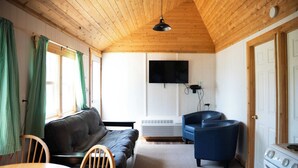A cozy room with a wooden ceiling, a black sofa, a blue armchair, a dining table with chairs, and a white stove.