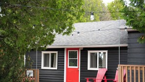 A small black house with a red door, two windows, and a porch with red chairs.