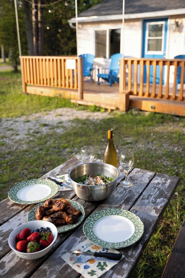 A wooden picnic table with plates, a bowl of strawberries, a bottle of wine, and a metal bowl with food. - Cozy 2‑Bed Waterfront Cabin | Near Bruce Peninsula (Tobermory)