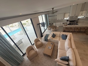 A modern living room with a sofa, armchairs, and a coffee table. A kitchen with white cabinets and a dining area with a table and chairs is visible. Large windows offer a view of an outdoor pool area.