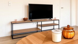 A modern living room with a flat-screen TV on a black metal stand, a wooden coffee table, a candle, and a jar on the table. - Central new-build apartment in Siegen (Siegen)