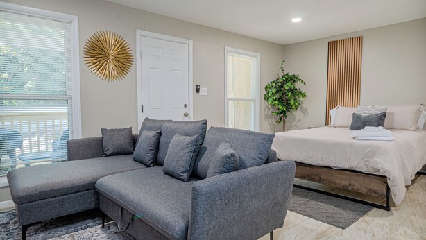 A modern bedroom with a grey sectional sofa, a bed with a wooden frame, a decorative wall piece, and a potted plant. - The Travelers Studio Columbus (Columbus)