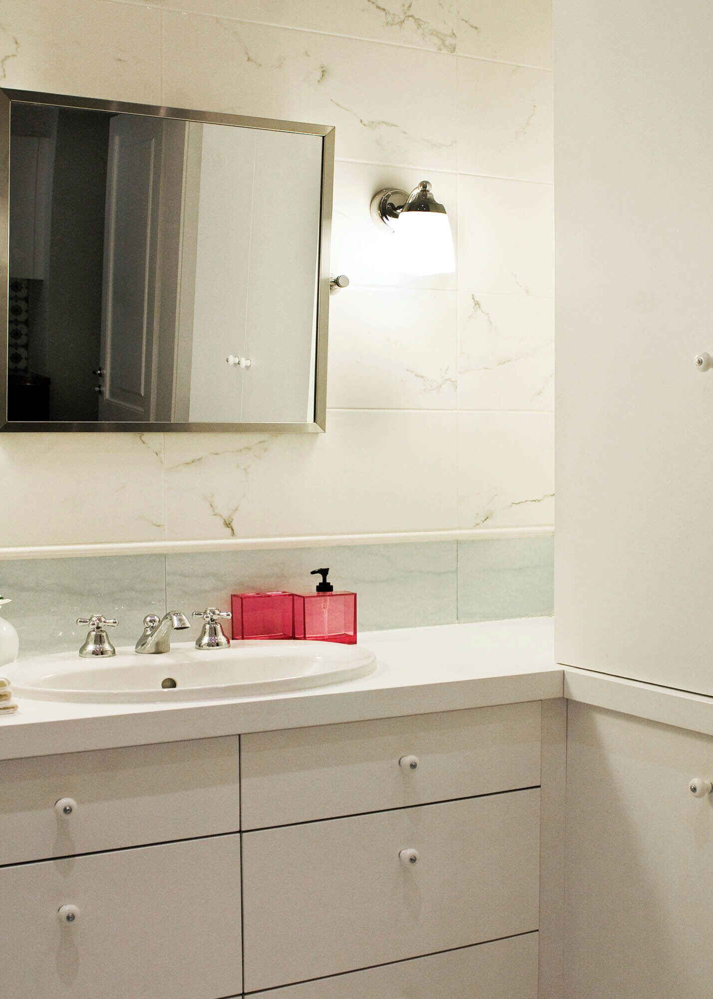 A bathroom with a marble-tiled wall, a white sink with three faucets, a red soap dispenser, and a mirror with a light fixture.