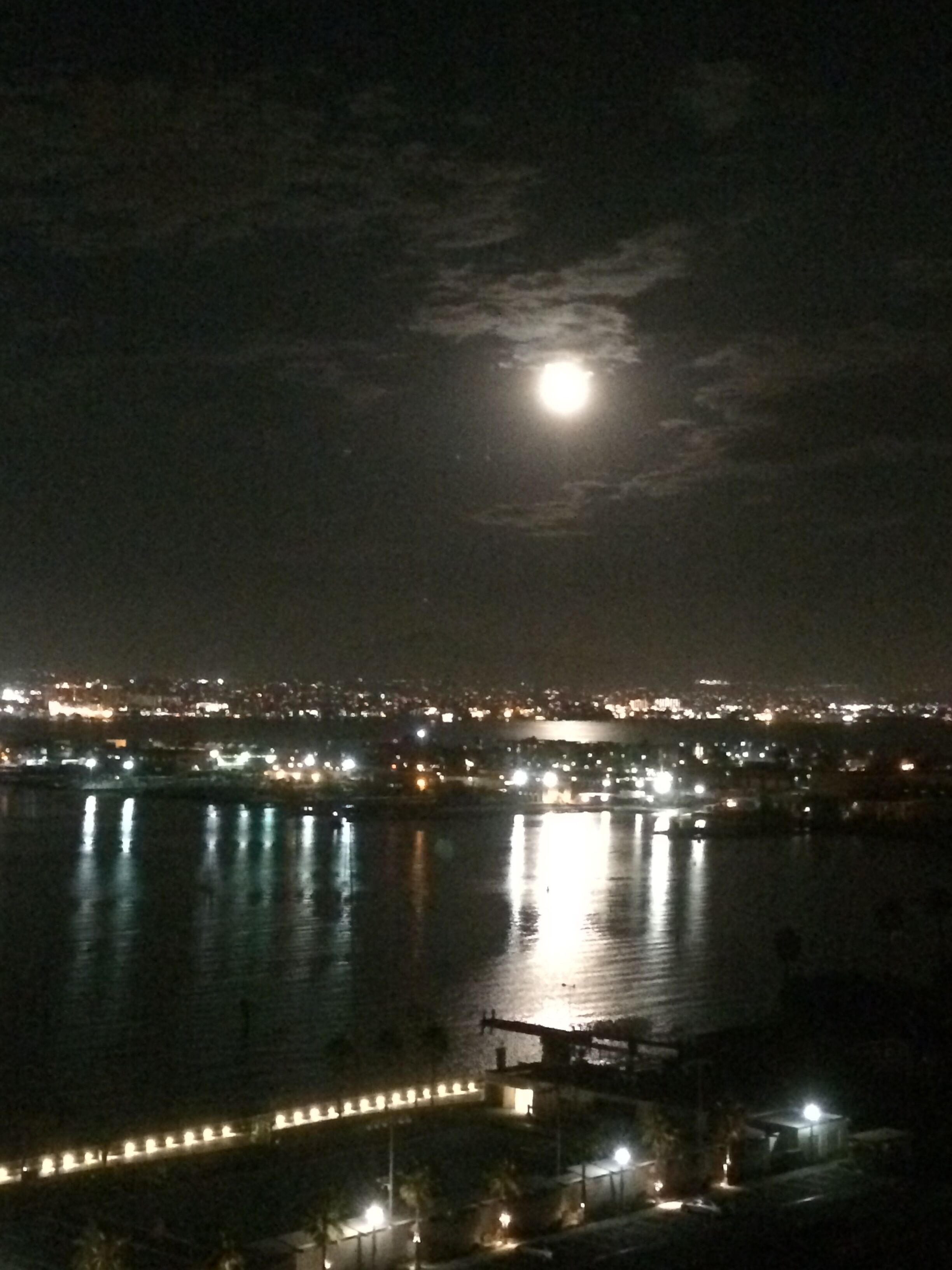 Une vue nocturne d’une ville, avec une pleine lune bien visible, une étendue d’eau reflétant les lumières et, au loin, le profil de la ville.