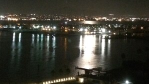 A nighttime view of a cityscape with a prominent full moon, a body of water reflecting lights, and a distant city skyline.