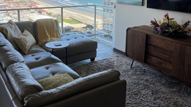 A modern living room with a sofa, a small round table, and a wooden cabinet with a floral arrangement. A large TV is mounted on the wall, and a balcony overlooks a beach and ocean view.