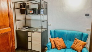 A compact room with a kitchenette, a blue sofa with orange cushions, and a wooden wardrobe.