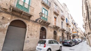 A narrow street lined with historic buildings, featuring balconies with metal railings, a row of parked cars, and a closed shop with a shuttered door. - Romano Apartment - Trapani (Trapani)