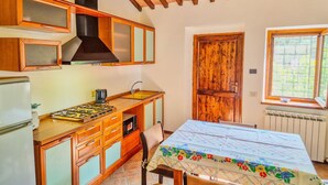 A compact kitchen with wooden cabinets, a gas stove, and a dining table with a floral tablecloth. - Monte Oliveto House - Apartment 3 (Bettona)