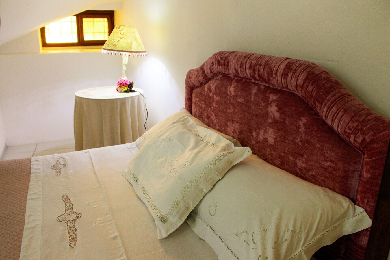 Ein Schlafzimmer mit Bett, Kissen, einem Nachttisch, einer Lampe und einem Fenster.