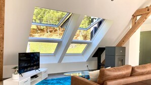 Un salon moderne avec un plafond en pente, de grandes fenêtres donnant sur une forêt, un téléviseur, un canapé en cuir marron et un tapis bleu.