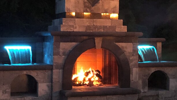 A stone-built outdoor fireplace with a fire burning inside, surrounded by stone arches and a decorative emblem above the fire. - Spacious 5-bedroom house in nice Winfield with WiFi, Pool and Hot tub (Winfield)