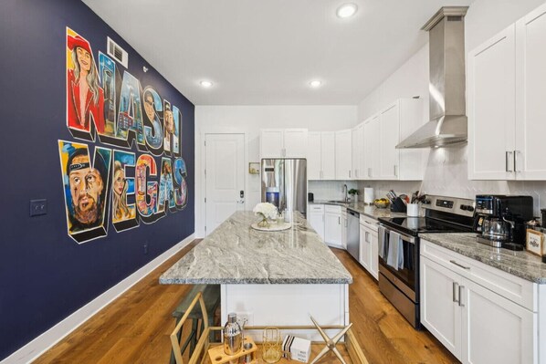 A modern kitchen with white cabinets, a marble countertop, and a central island. There is a mural on the wall depicting various people and the text "NASH VEGAS".