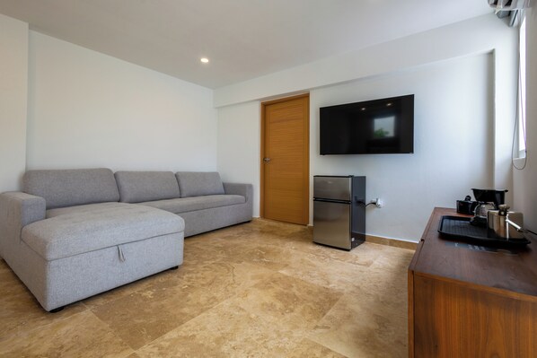 A modern living room with a grey sectional sofa, a wooden door, a flat-screen TV, a refrigerator, and a wooden coffee table with a kettle. - Anchors Hotel (Fajardo)