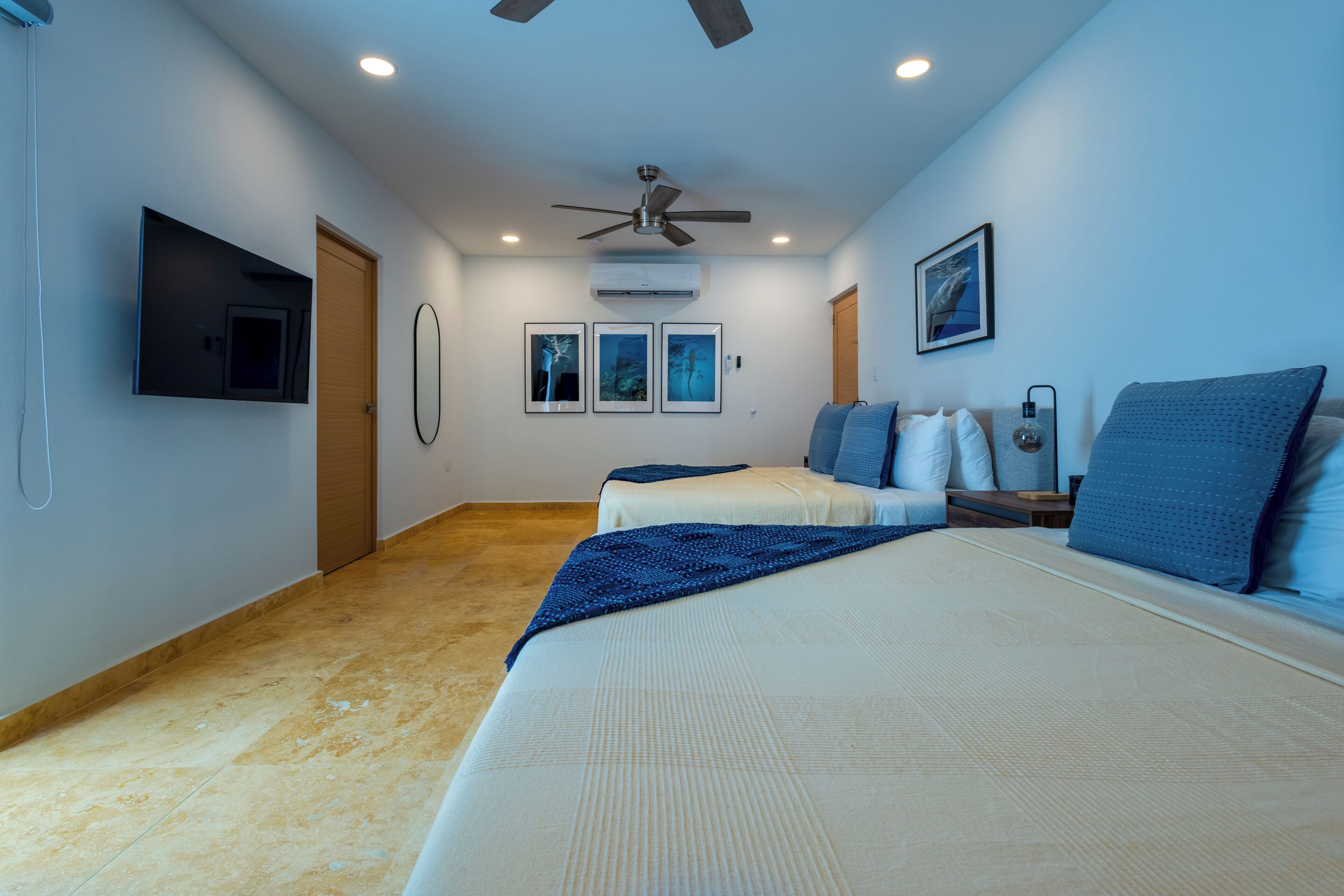 Une chambre avec deux lits, un ventilateur de plafond, un téléviseur fixé au mur et des tableaux encadrés accrochés au mur.