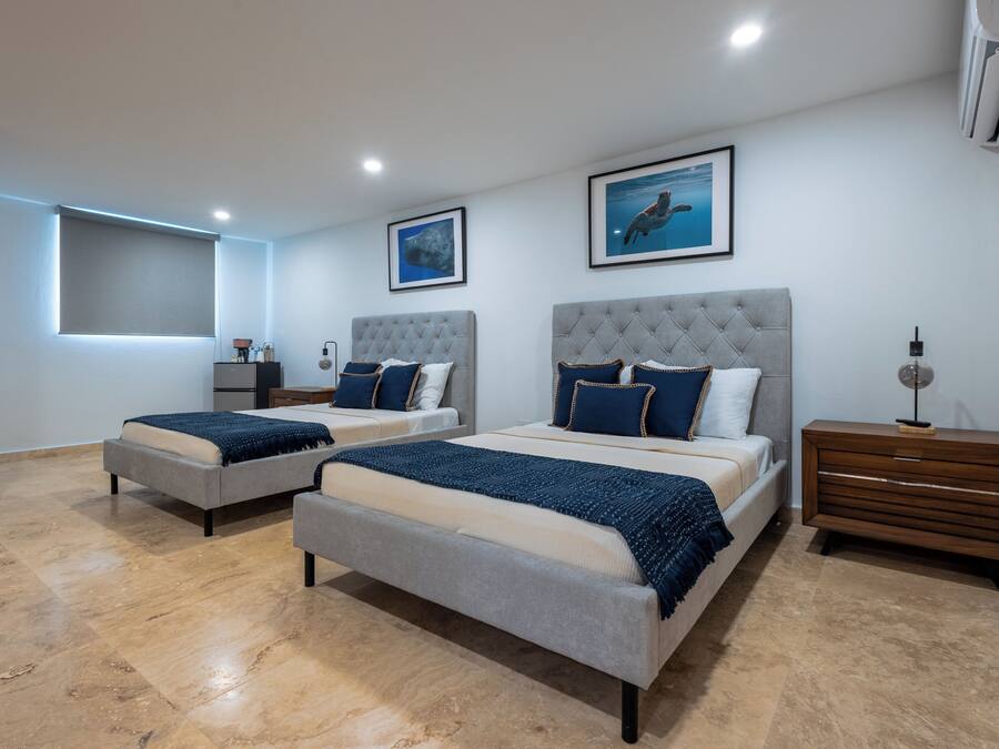 A modern bedroom with two beds, a large window with blinds, and a nightstand with a lamp.