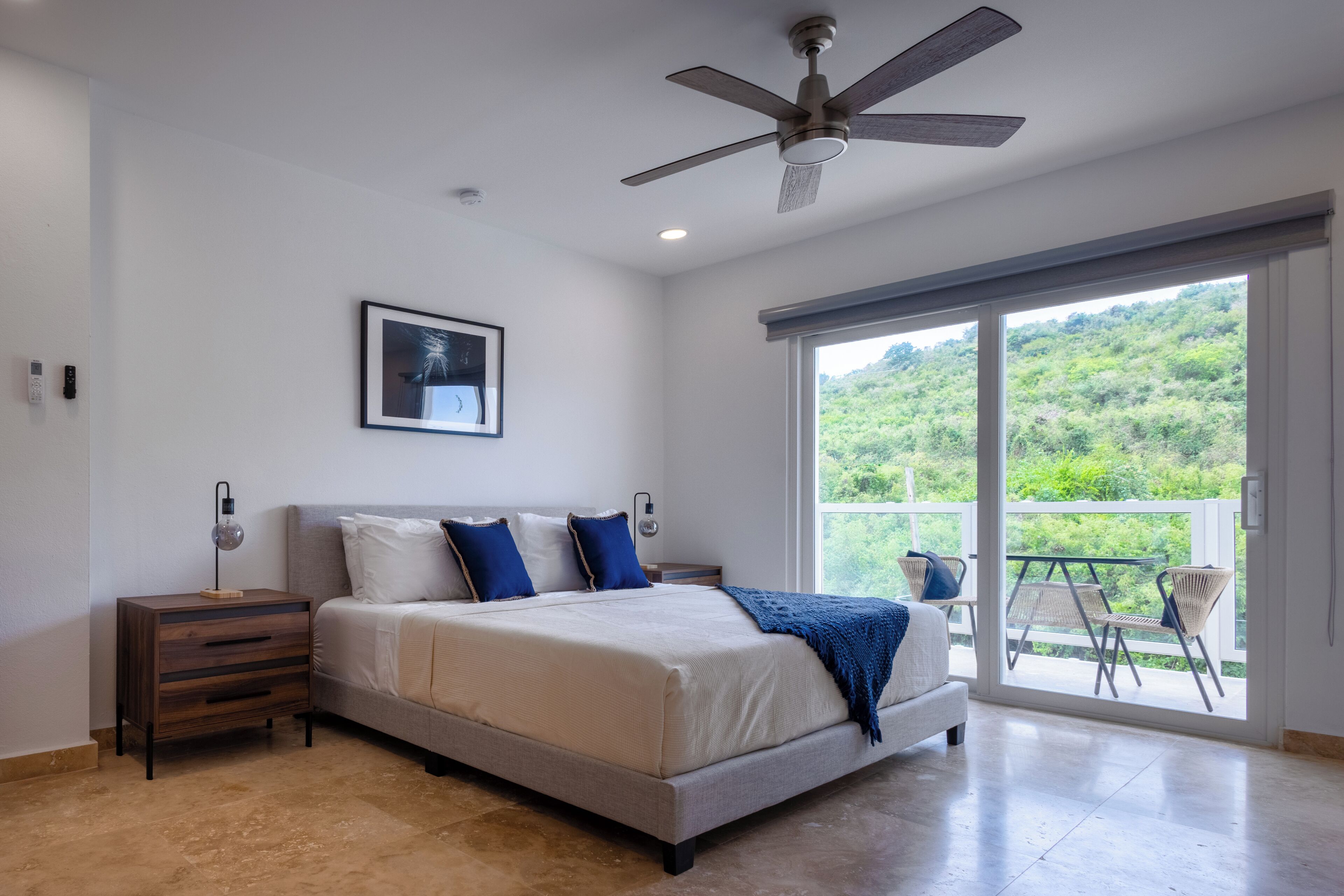 A bedroom with a bed, a nightstand, a ceiling fan, a window with a view of greenery, and a balcony with chairs.