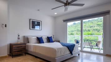 A bedroom with a bed, a nightstand, a ceiling fan, a window with a view of greenery, and a balcony with chairs.