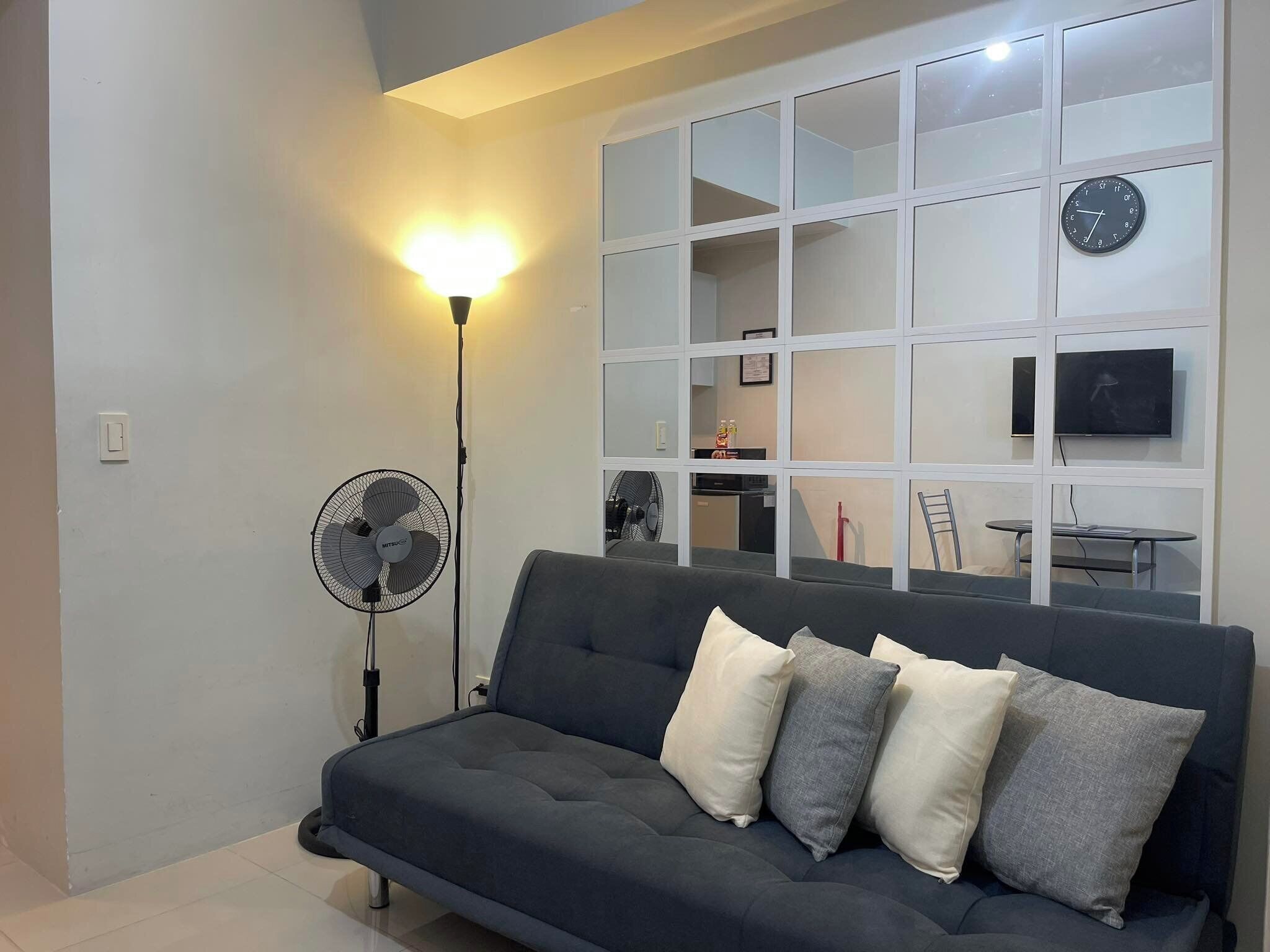 Un salon moderne avec un canapé foncé, de nombreux coussins, un ventilateur sur pied et un téléviseur fixé au mur.