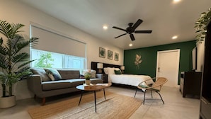 Un salon moderne avec un canapé, une table basse et un fauteuil, un ventilateur de plafond et un lit, le tout dans une pièce dont un mur est peint en vert et qui possède une porte.
