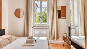 Een slaapkamer met een bed, een nachtkastje met een plant, een raam met gordijnen en een spiegel aan de muur.