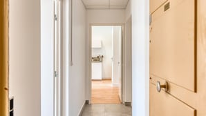 Een smalle gang met een houten deur aan de rechterkant en een witte deur aan de linkerkant, die uitkomt in een keukenruimte met een aanrecht en kastjes.