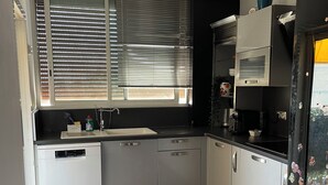 Una cocina compacta con gabinetes blancos, lavavajillas, un cubo de basura azul y una ventana con persianas.