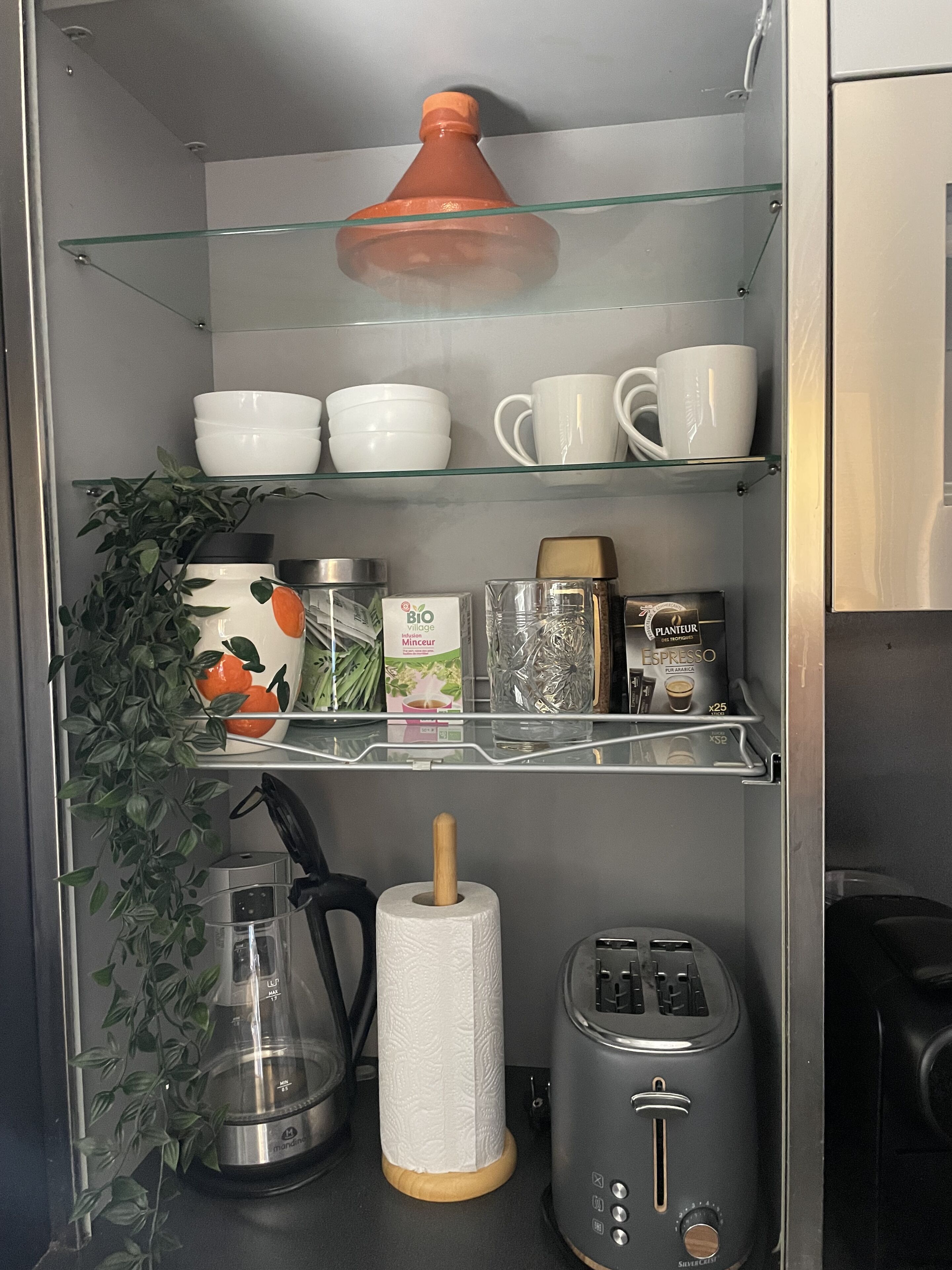 A kitchen with a coffee maker, a toaster, a kettle, and a shelf with cups and bowls.