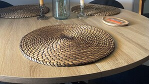Una mesa redonda de madera con individuales tejidos, un florero de vidrio con flores y velas. Un reloj de pared grande está colgado sobre la mesa.