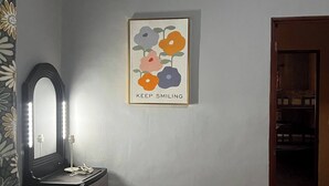 A room with a black dresser, a mirror with lights, a wall-mounted air conditioner, and a framed floral artwork with the text "KEEP SMILING".
