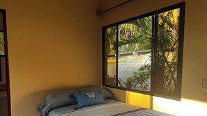 A bedroom with a bed, a window with a view of palm trees, and a wall-mounted light fixture.