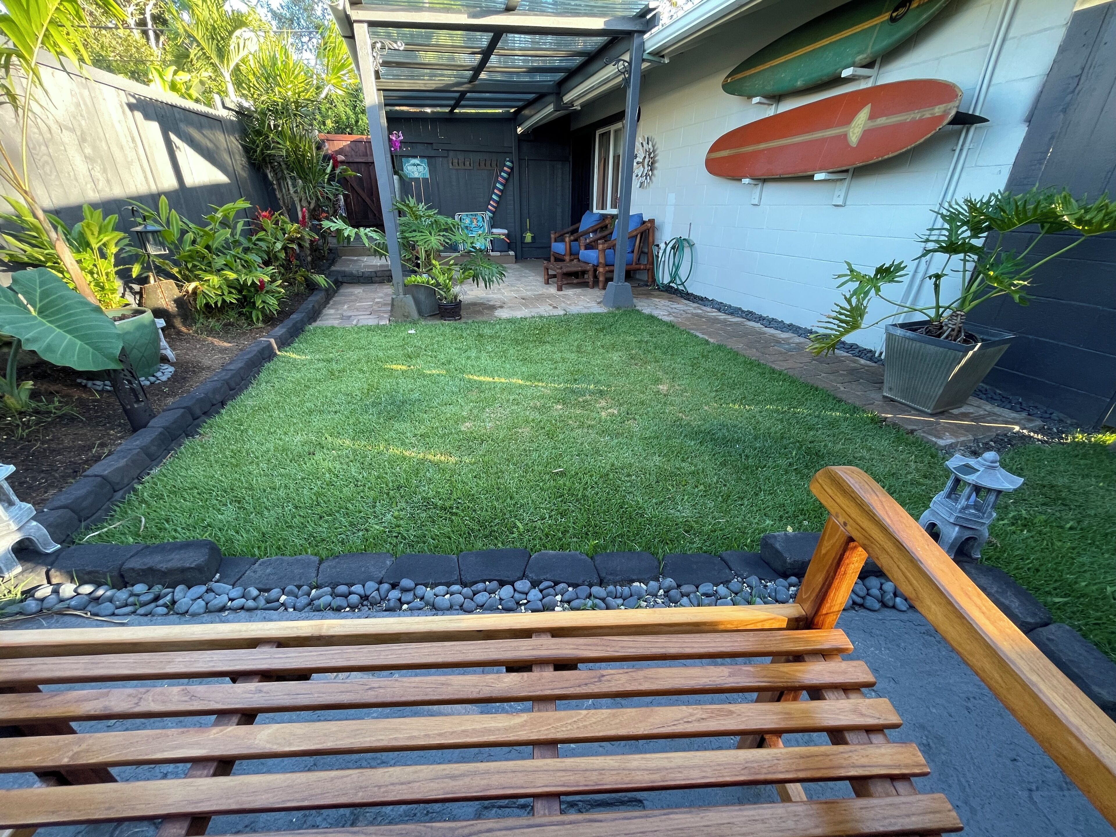A backyard with a grassy lawn, a wooden bench, and a surfboard on the wall.