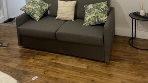 Ein graues Sofa mit drei Kissen, ein Wandbild mit Blättern und ein kleiner Tisch mit Vase und Schilferdiffusor.