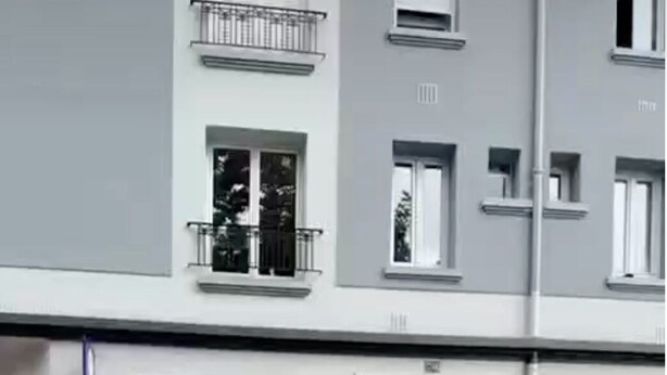 Ein mehrstöckiges Gebäude mit grauem Mauerwerk, einem Metallgitter am Eingang und einer kleinen Gartenanlage mit Pflanzen.