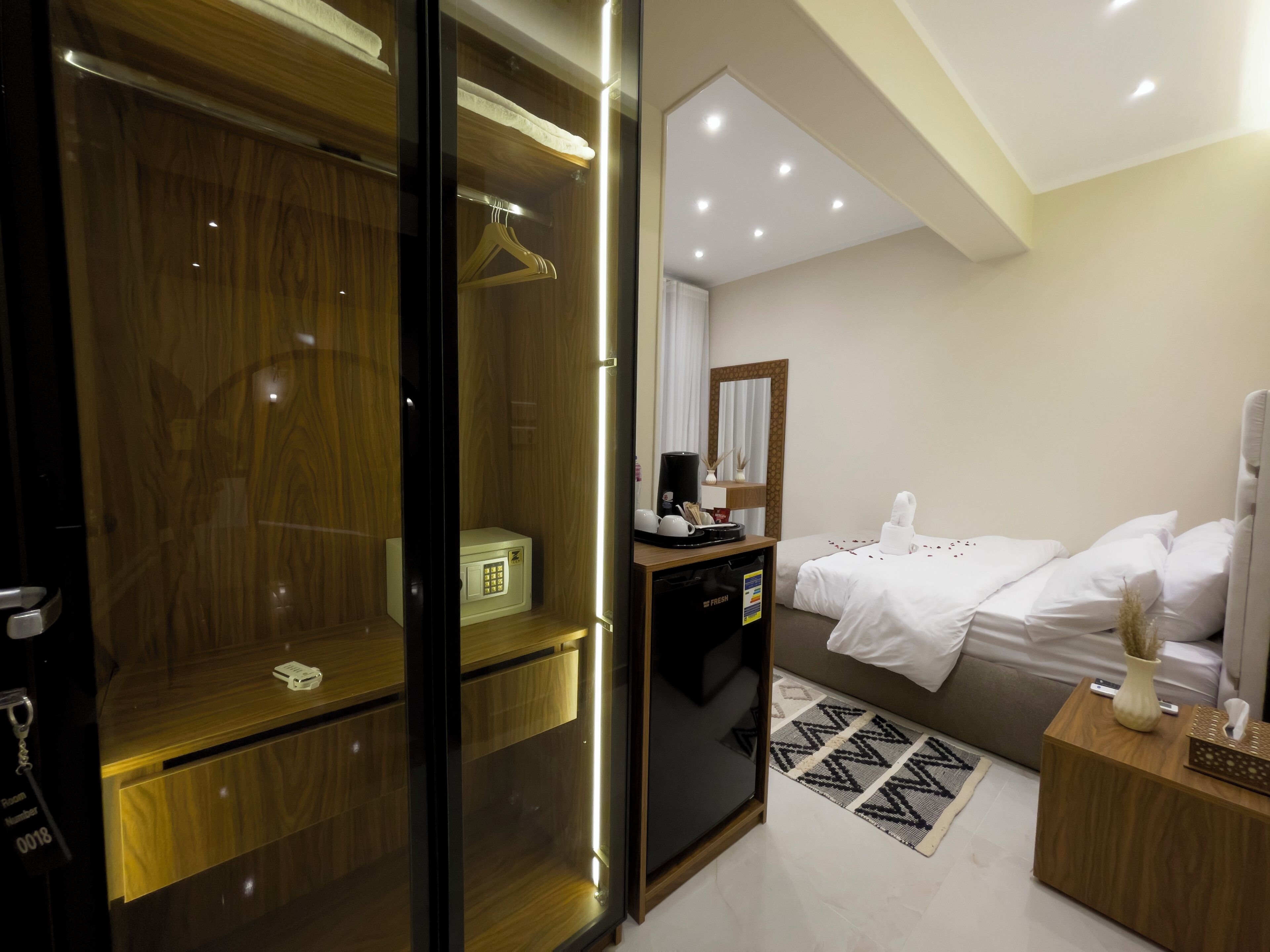A modern hotel room with a walk-in closet, a bed with white linens, a bedside table with a vase, and a small desk with a coffee maker.