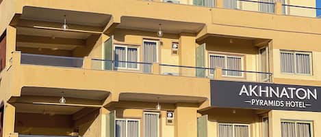 A multi-story hotel with balconies, a sign reading 'AKHNATON Pyramids Hotel,' and outdoor umbrellas.