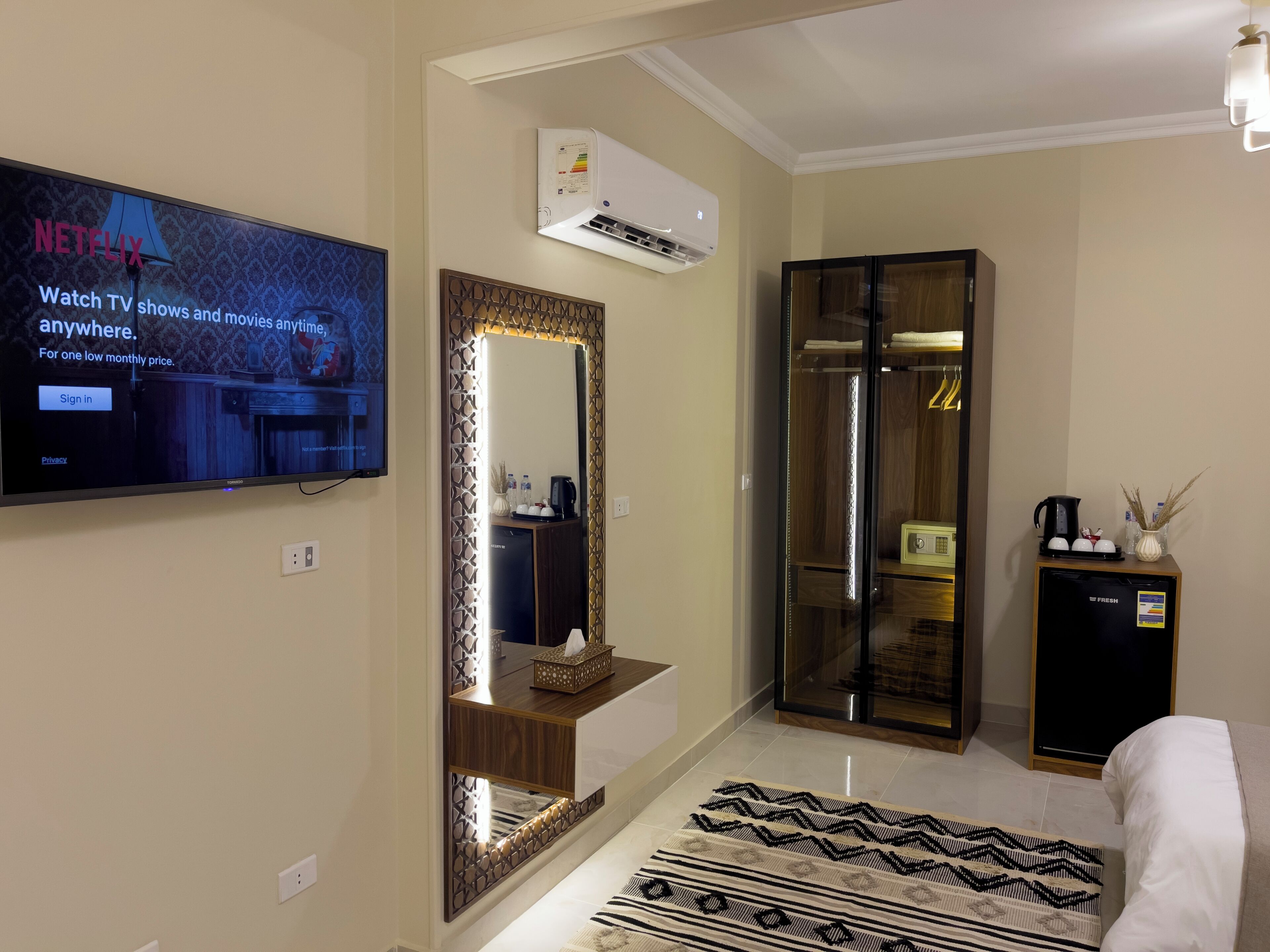 Ein Hotelzimmer mit einem Fernseher, der ein Netflix-Logo zeigt, einem Spiegel, einem Kleiderschrank, einem Nachttisch mit einem Radio und einem gemusterten Teppich.