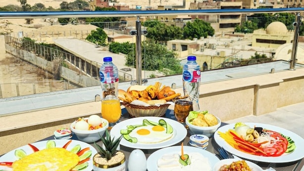 A table with various breakfast dishes and drinks, overlooking a cityscape with pyramids in the background. - Akhnaton Pyramids View (Giza)