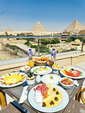 Une table dressée avec divers plats et boissons pour le petit-déjeuner, offrant une vue sur un paysage urbain avec des pyramides en arrière-plan.
