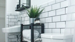 Un baño con inodoro blanco, lavamanos, espejo y ducha. Hay una planta sobre el lavamanos y toallas en el estante.