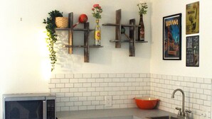 Una cocina con microondas, lavamanos y salpicadero de azulejos blancos. Hay estantes con adornos y una planta en maceta.